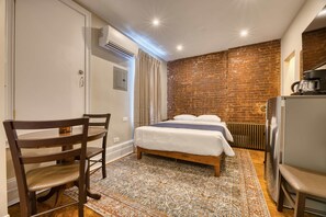 A bedroom with a brick wall, a bed with white bedding, a wooden chair, a small table, and a radiator.