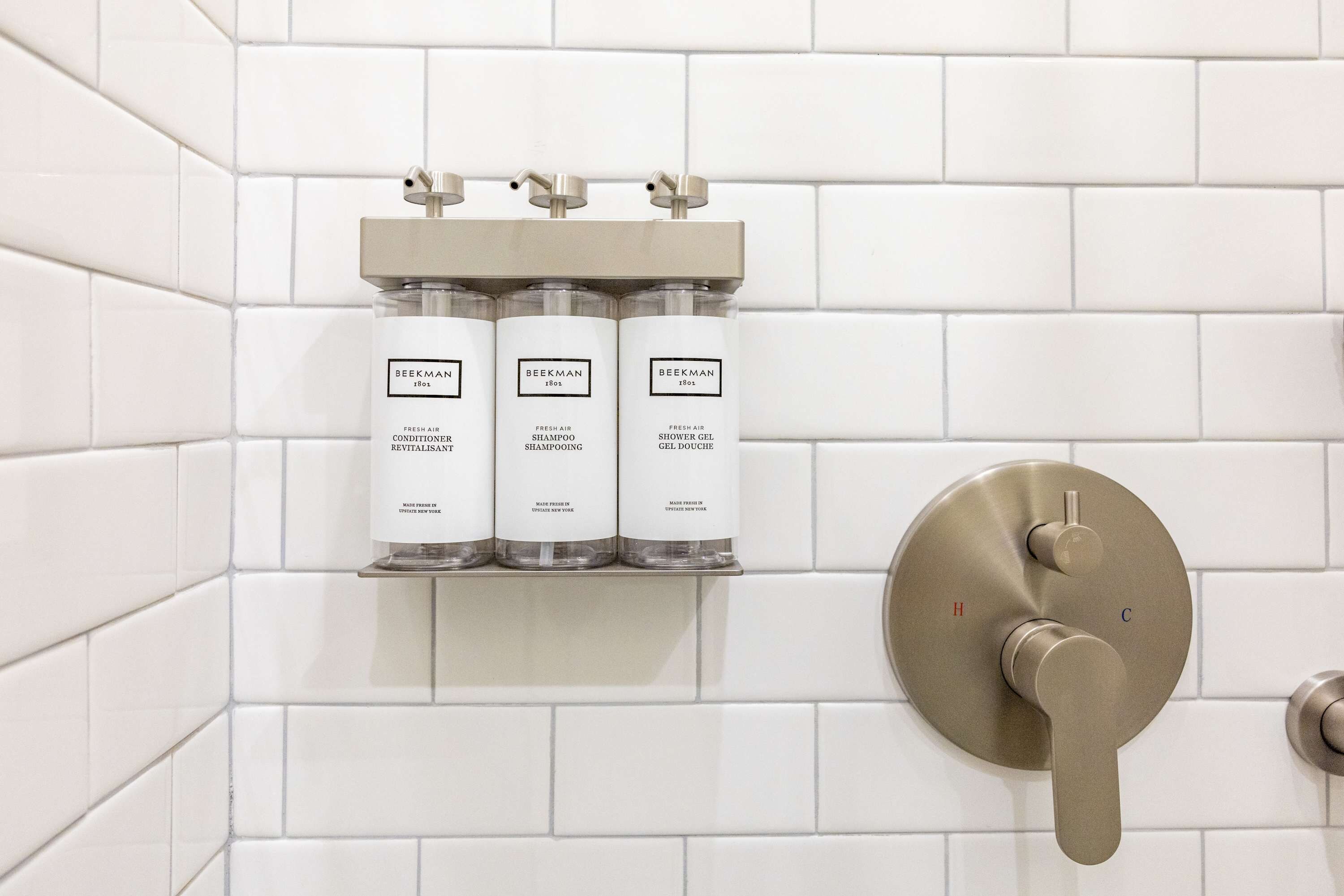 Ein Badezimmer mit weißen Fliesenwänden, einem Regal mit drei Produkten von Beekman und einem modernen Wasserhahn.
