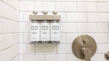 Ein Badezimmer mit weißen Fliesenwänden, einem Regal mit drei Produkten von Beekman und einem modernen Wasserhahn.