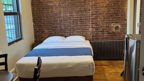 A hotel room with a brick wall, a bed with white bedding, a wooden floor, a chair, and a refrigerator.