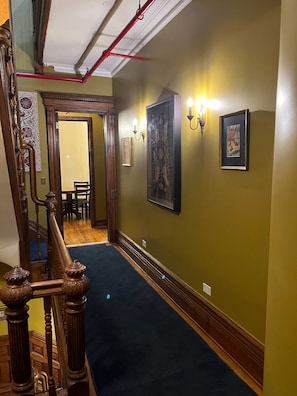 Un pasillo con escalera de madera, alfombra azul y lámparas fijadas a la pared.