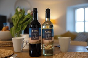Zwei Weinflaschen, beschriftet mit 'Rotwein' und 'Weißwein', auf einem Tisch. Im Hintergrund steht eine Pflanze und eine Tasse.