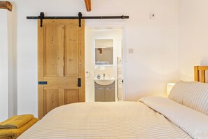 A bedroom with a bed, a wooden door, a bathroom with a sink, and a mirror. - Rose Cottage - Sea Views, Hot Tub, Log Burner (Pendine)