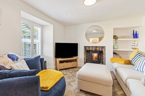 Een woonkamer met een televisie, open haard, bank, poef en een raam met jaloezieën.