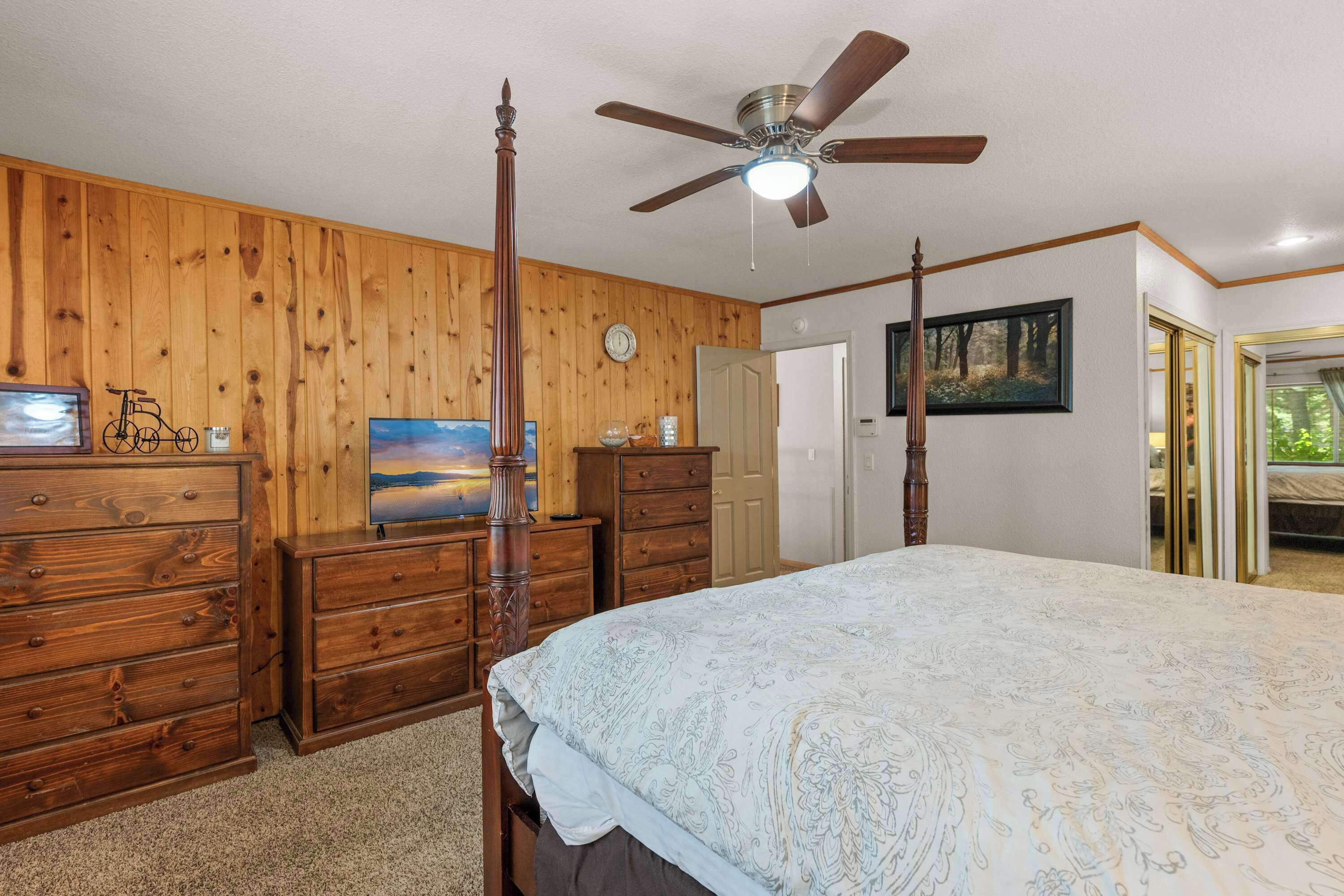 Ein Schlafzimmer mit einer Holzvertäfelung, einem Deckenventilator, einem Bett mit gemustertem Überwurf, einem Schminktisch und einem Gemälde.