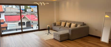 Una sala de estar moderna con un sofá modular, un ventilador de pie y vistas a edificios a través de amplias ventanas.
