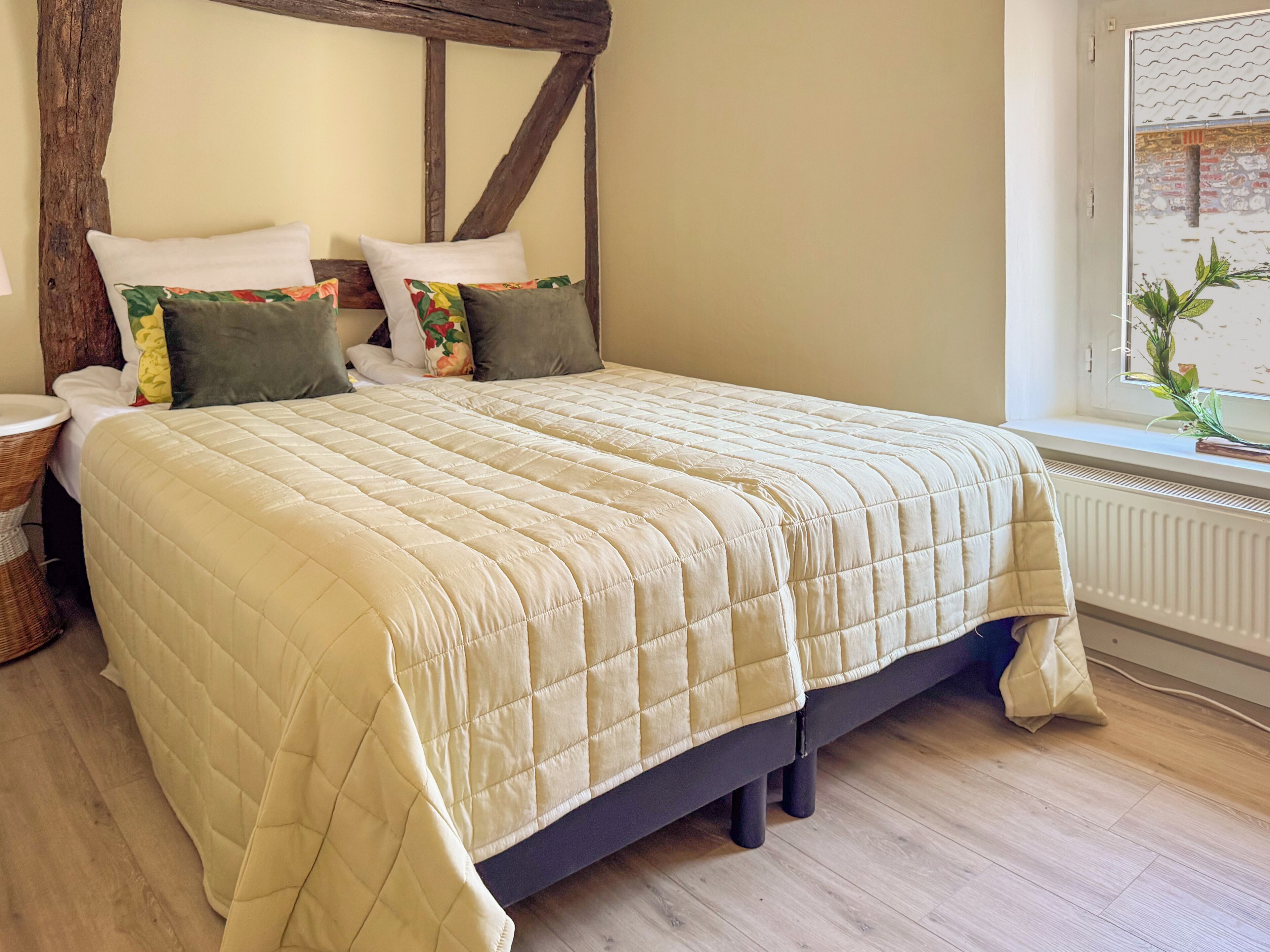 A bed with a quilted bedspread, two pillows, a nightstand, and a window with a view of greenery.