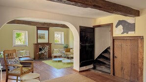 Una habitación con vigas de madera, chimenea, sofá, mesa de comedor y escalera.