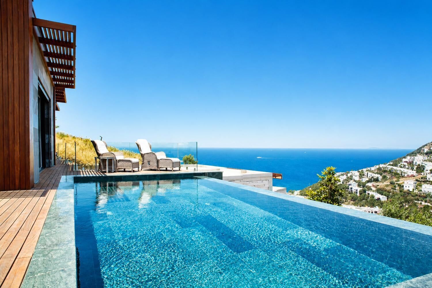 A modern infinity pool with clear blue water, wooden deck, and lounge chairs, overlooking a coastal town and the sea.