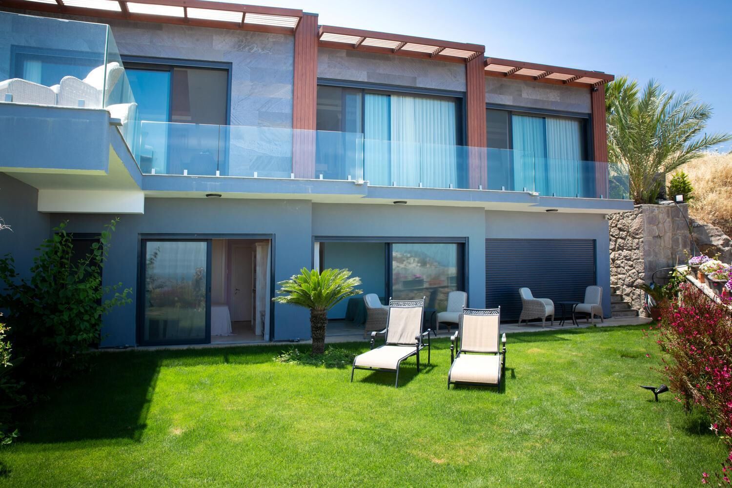 Une maison moderne à deux étages, dotée d’un balcon, d’une terrasse avec des chaises et d’une pelouse bien entretenue.