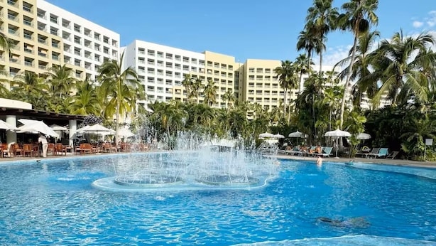Ein Pool mit einer Fontäne, umgeben von Palmen und Hotelgebäuden.