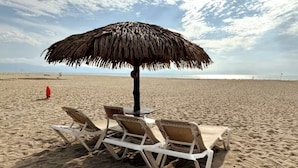 Ein Strand mit einem Reetdachschirm, zwei Liegestühlen und einem kleinen Tisch.