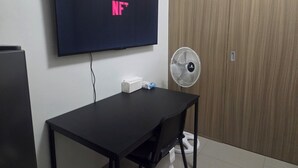Ein Zimmer mit Fernseher, der 'NF' anzeigt, einem Schreibtisch, einem Stuhl, einem Standventilator und einem Kleiderschrank.