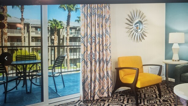 A room with a view of a building with balconies, a patterned curtain, a sunburst wall decoration, a lamp, a green sofa, and a table with a basket of apples.