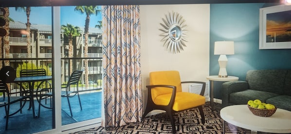 A room with a view of a building with balconies, a patterned curtain, a sunburst wall decoration, a lamp, a green sofa, and a table with a basket of apples.
