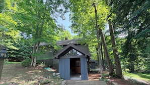 A wooden cabin with a porch, surrounded by trees and a stone pathway.