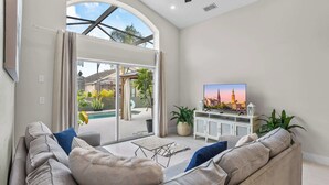 A living room with a sofa, TV, and a view of an outdoor pool area. - Fire Pit Pool Retreat for Groups (Kissimmee)