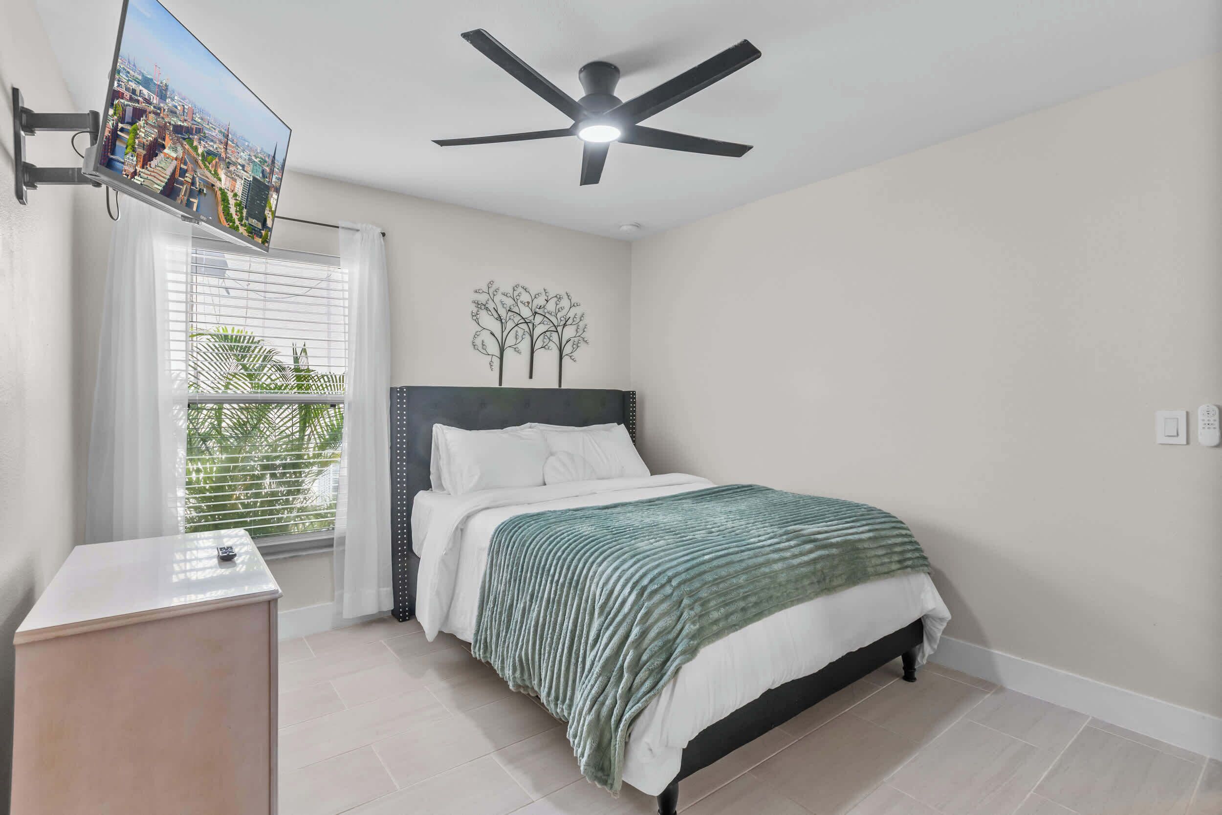Une chambre à coucher avec un lit, un ventilateur de plafond, un téléviseur fixé au mur et une fenêtre avec des stores.