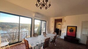 A dining area with a table set for four, large windows with a view of a river and trees, and a chandelier. - 4-bedroom house, sleeps 8, lake view. (Château-la-Vallière)