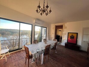 A dining area with a table set for four, large windows with a view of a river and trees, and a chandelier. - 4-bedroom house, sleeps 8, lake view. (Château-la-Vallière)