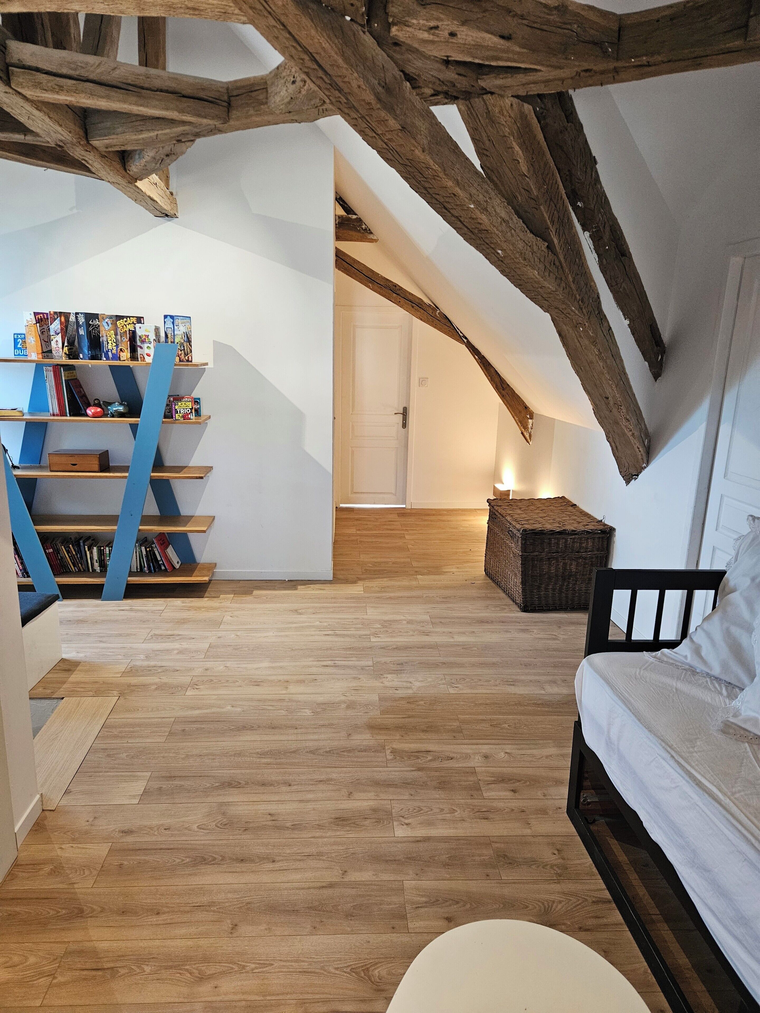 An attic bedroom with exposed wooden beams, a white bed, a bookshelf, and a wooden floor.