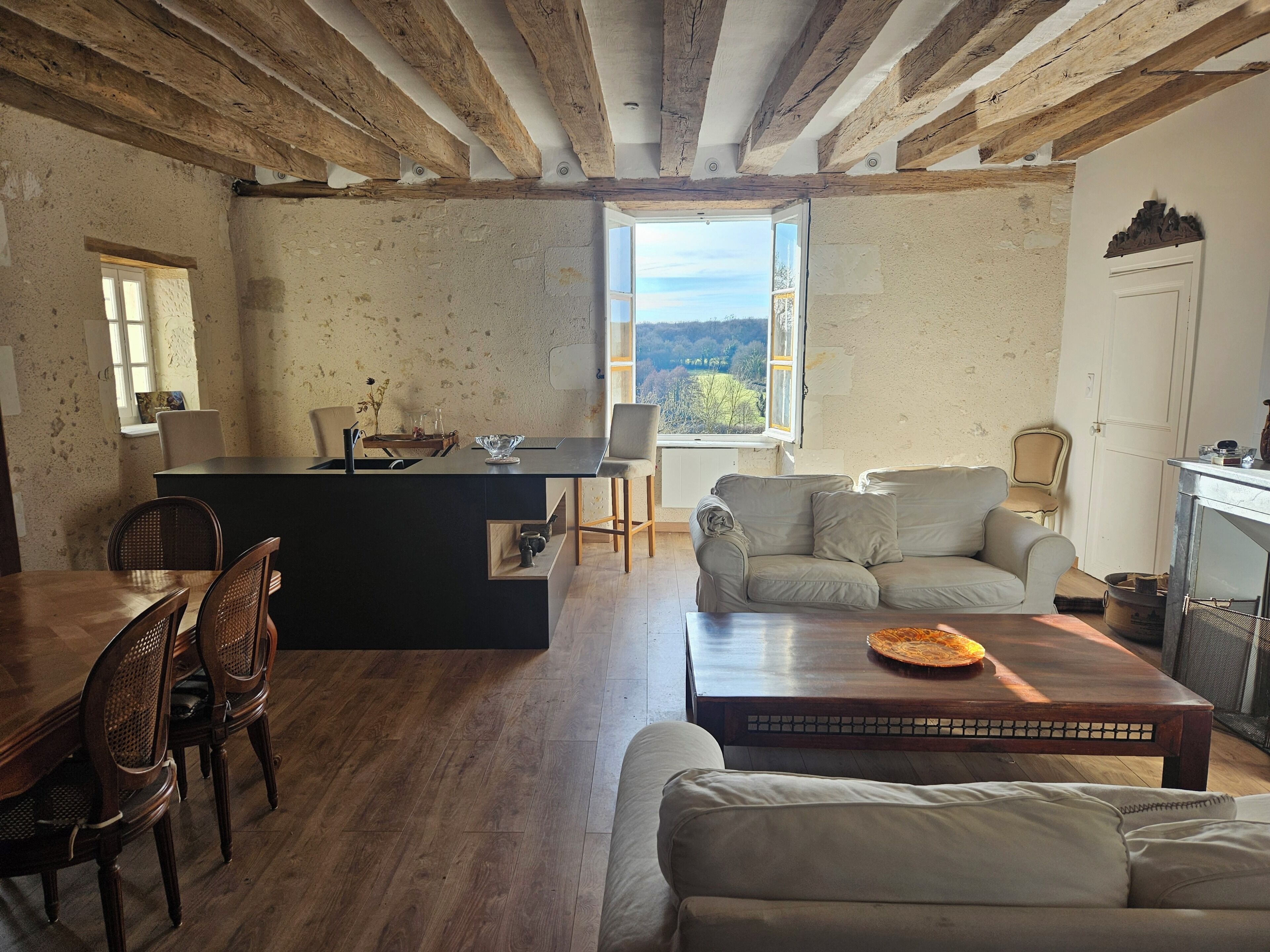 Een rustiek interieur met zichtbare houten balken, een eethoek met een houten tafel en stoelen, een bank en een uitzicht door een raam.