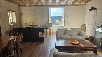 Een rustiek interieur met zichtbare houten balken, een eethoek met een houten tafel en stoelen, een bank en een uitzicht door een raam.