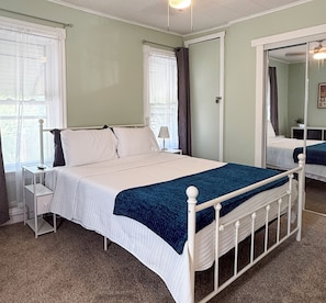 A neatly arranged bedroom with a single bed, a bedside table, a nightstand, a mirror, and a window with curtains.