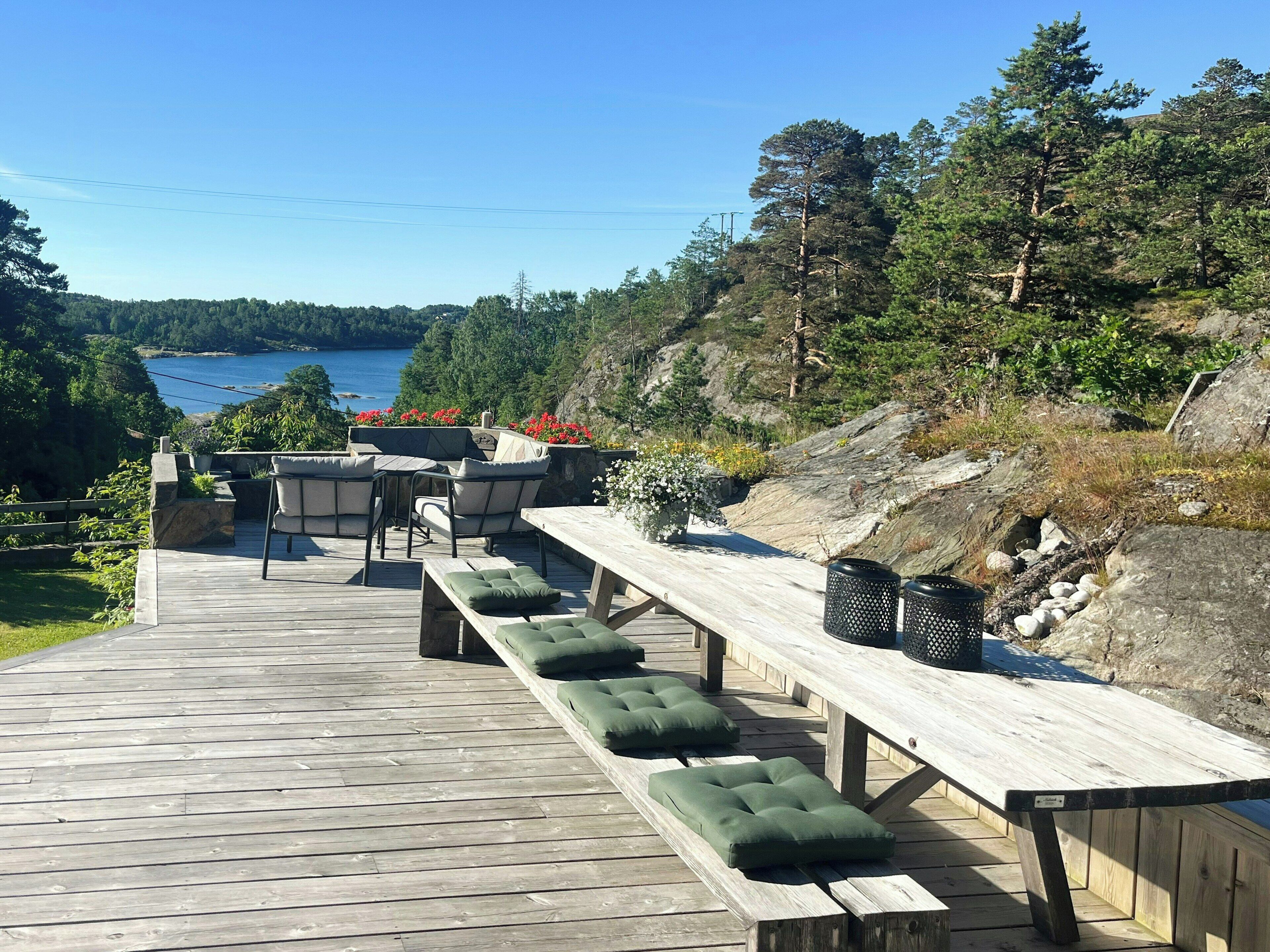 Una terraza de madera con mobiliario exterior, vista a un lago y vegetación circundante.