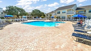 Ein Pool mit Liegestühlen und Sonnenschirmen, umgeben von einer Pflasterterrasse und einem weißen Zaun.