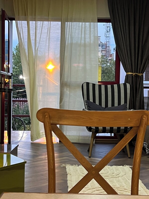 A room with a wooden chair, a striped sofa, a white rug, and a view through a window with curtains. - REZIDENCA 4R (Shkoder)