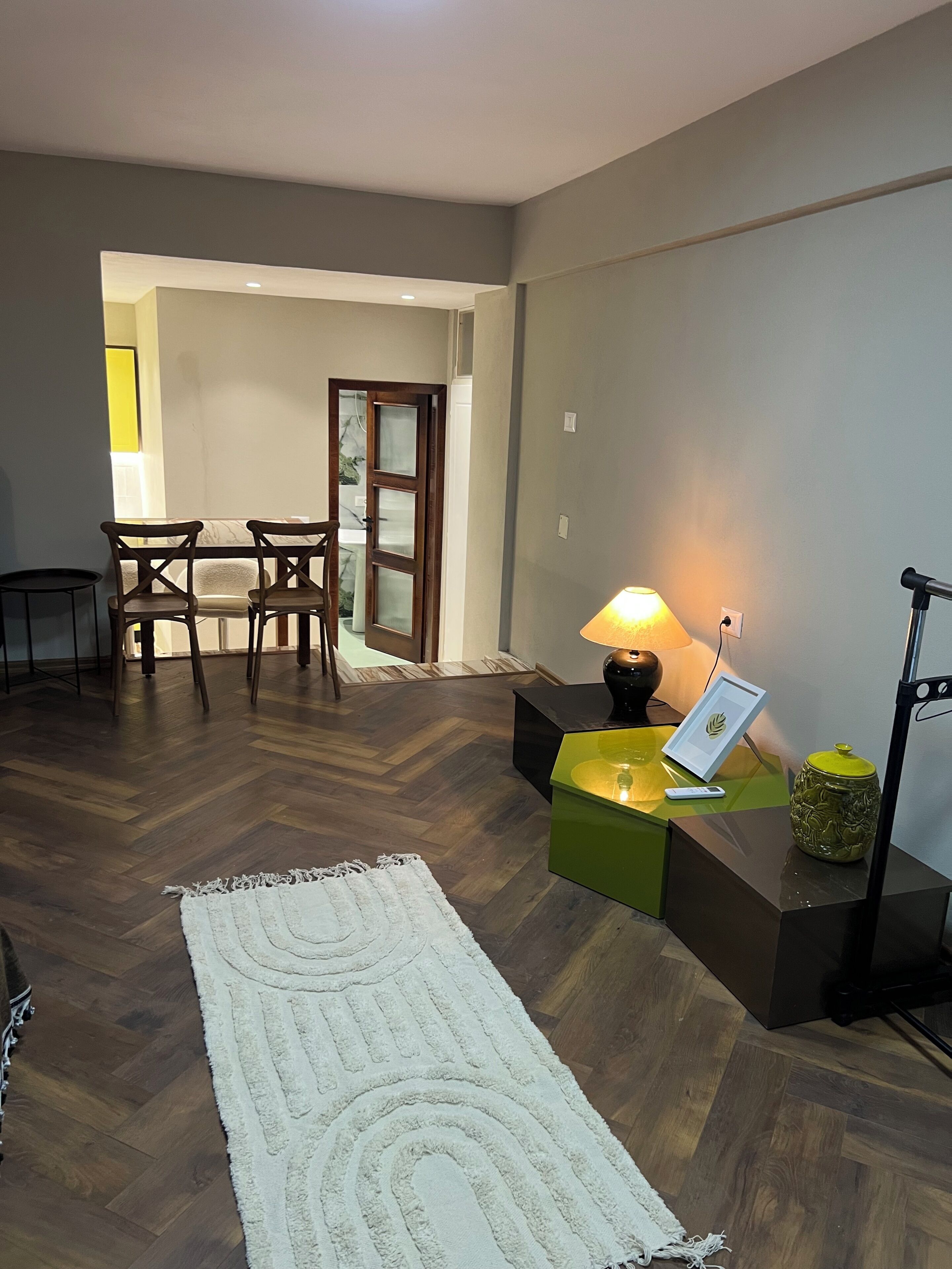 Un soggiorno con pavimento in legno, un tappeto bianco, un tavolino verde con una lampada e un quadro incorniciato.