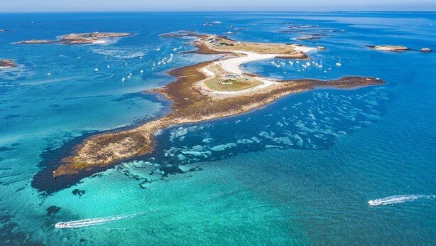 Vista aérea de uma área costeira com águas azul-claras, praias de areia e barcos espalhados.