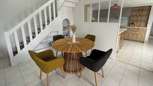 Um interior moderno com uma mesa de jantar redonda de madeira, duas cadeiras verdes e uma cadeira preta. Uma escada branca com corrimão leva a uma área de cozinha.