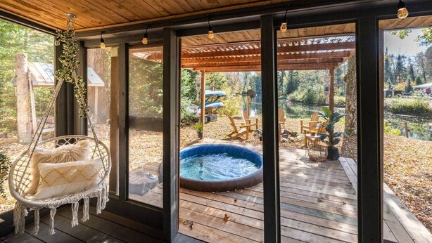 A wooden deck with a hanging chair, a hot tub, and outdoor furniture. - Hills Haven - Luxury Riverfront Cottage (Minden Hills)