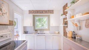 A modern kitchen with white cabinets, a stainless steel stove, a wooden sign above the sink, and a window with a view of greenery. - Hills Haven - Luxury Riverfront Cottage (Minden Hills)