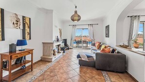 Ein Wohnzimmer mit einer Couch, einem Couchtisch, einem Fernseher und einem Kamin. Durch ein großes Fenster bietet sich ein Ausblick ins Freie.