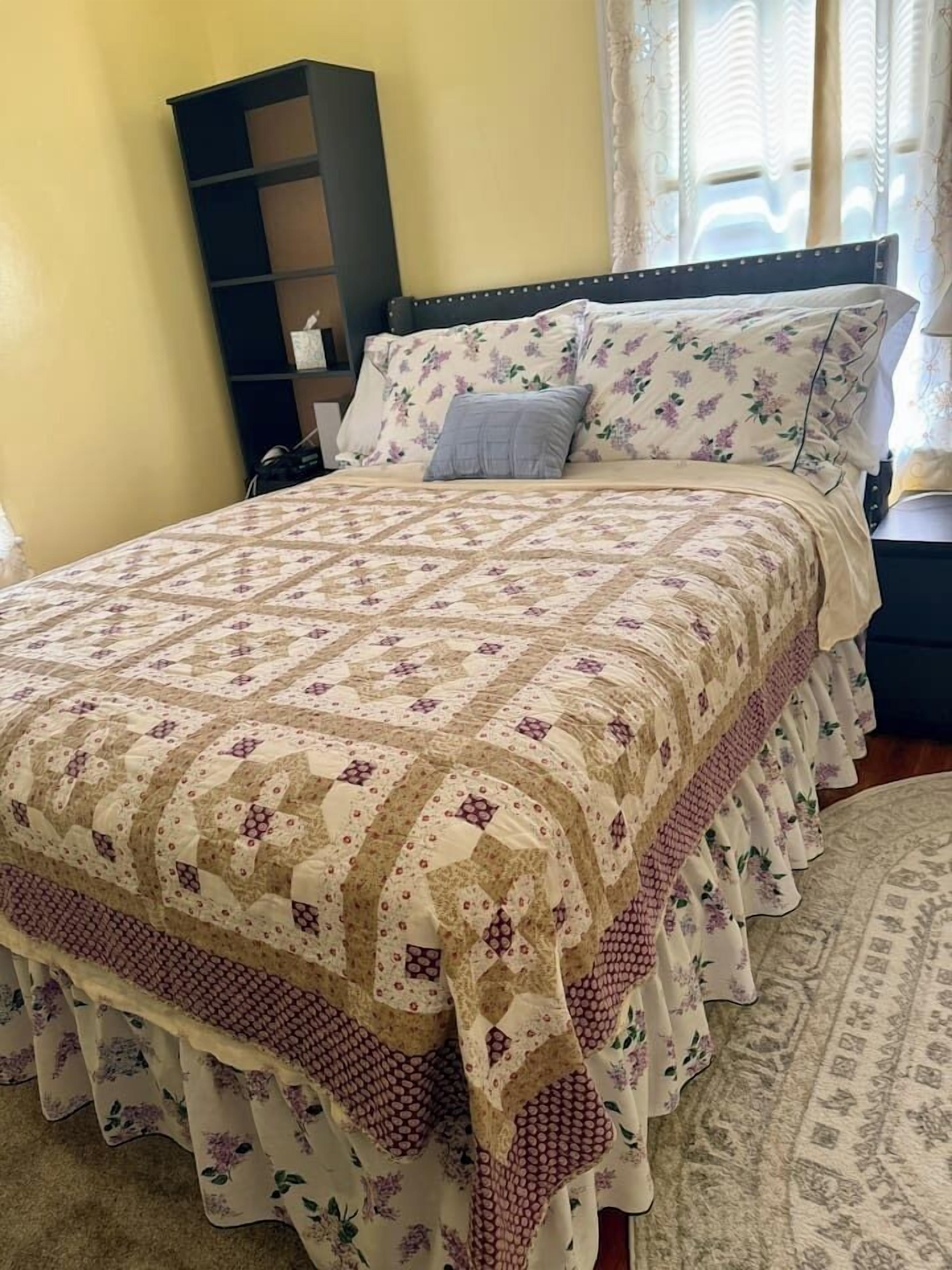 A bed with a floral-patterned bedspread and pillows, a dark-colored bookshelf, and a patterned rug on the floor.