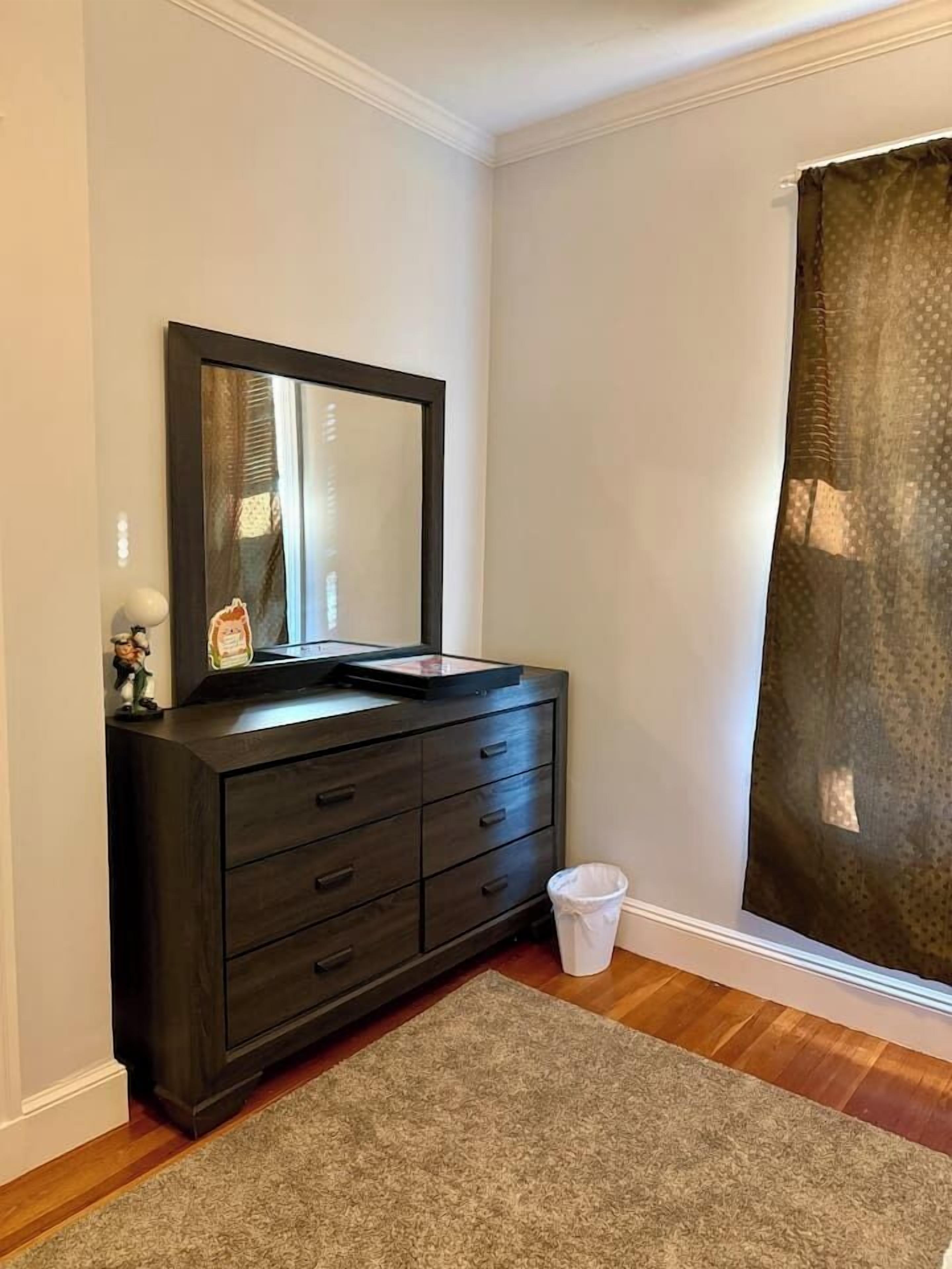Een kamer met een donker houten dressoir, een spiegel, een klein afvalbakje en een raam met gordijn.