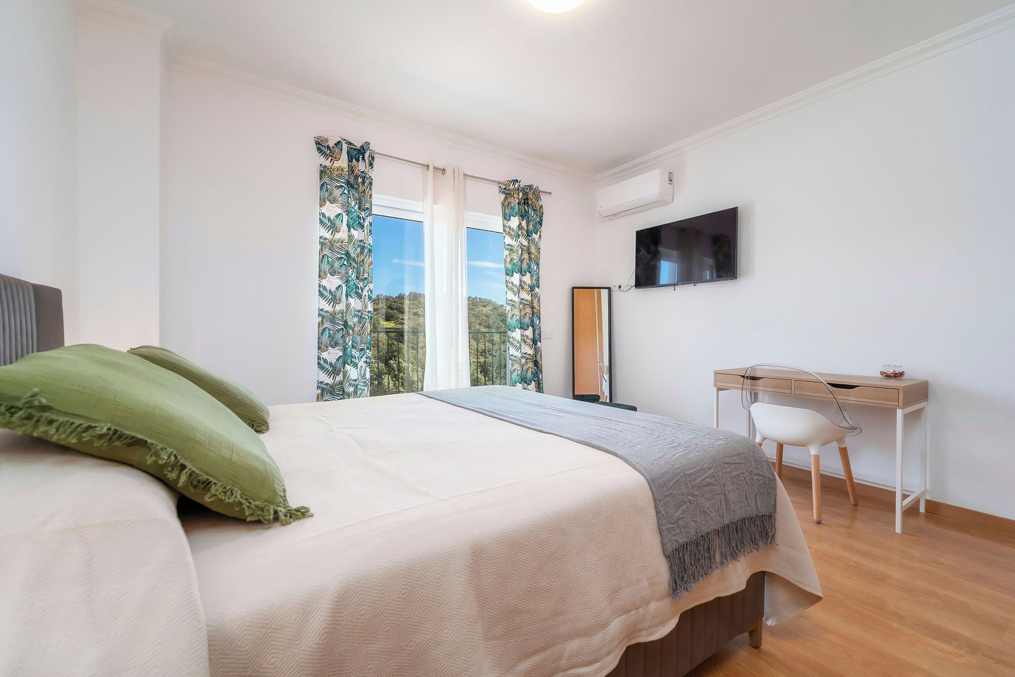 A bedroom with a bed, a desk, a chair, a TV, and a window with curtains.