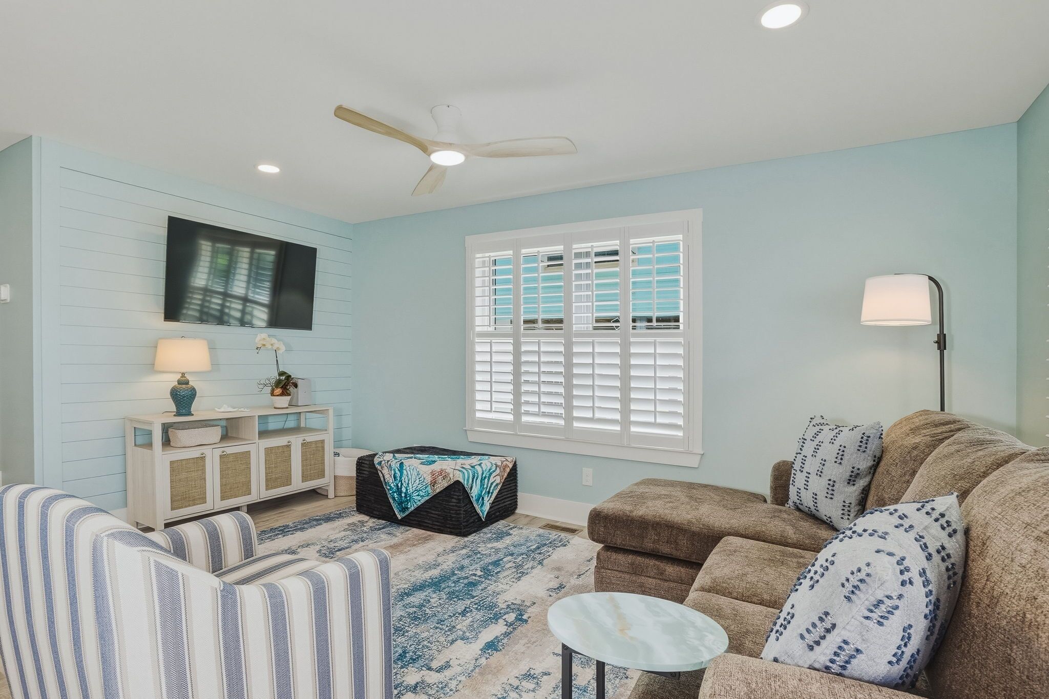 A living room with a sofa, a coffee table, a side table, a lamp, a television, a ceiling fan, and a window with shutters.