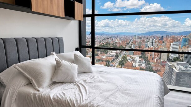 A bedroom with a bed, pillows, and a large window with a city view. - Apartment with incredible city view (Bogotá)
