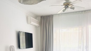 Un soggiorno moderno con televisione a schermo piatto, ventilatore a soffitto, tavolo da pranzo e un'ampia finestra con tende.