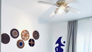 Camera d'albergo con un letto, due cuscini, una ventola a soffitto, una parete decorata con piatti, una lampada a parete e una finestra con tende.
