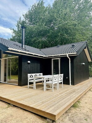 Ein modernes Haus mit dunkler Fassade, einem geneigten Dach und einer Holzterrasse mit Tisch und Stühlen.