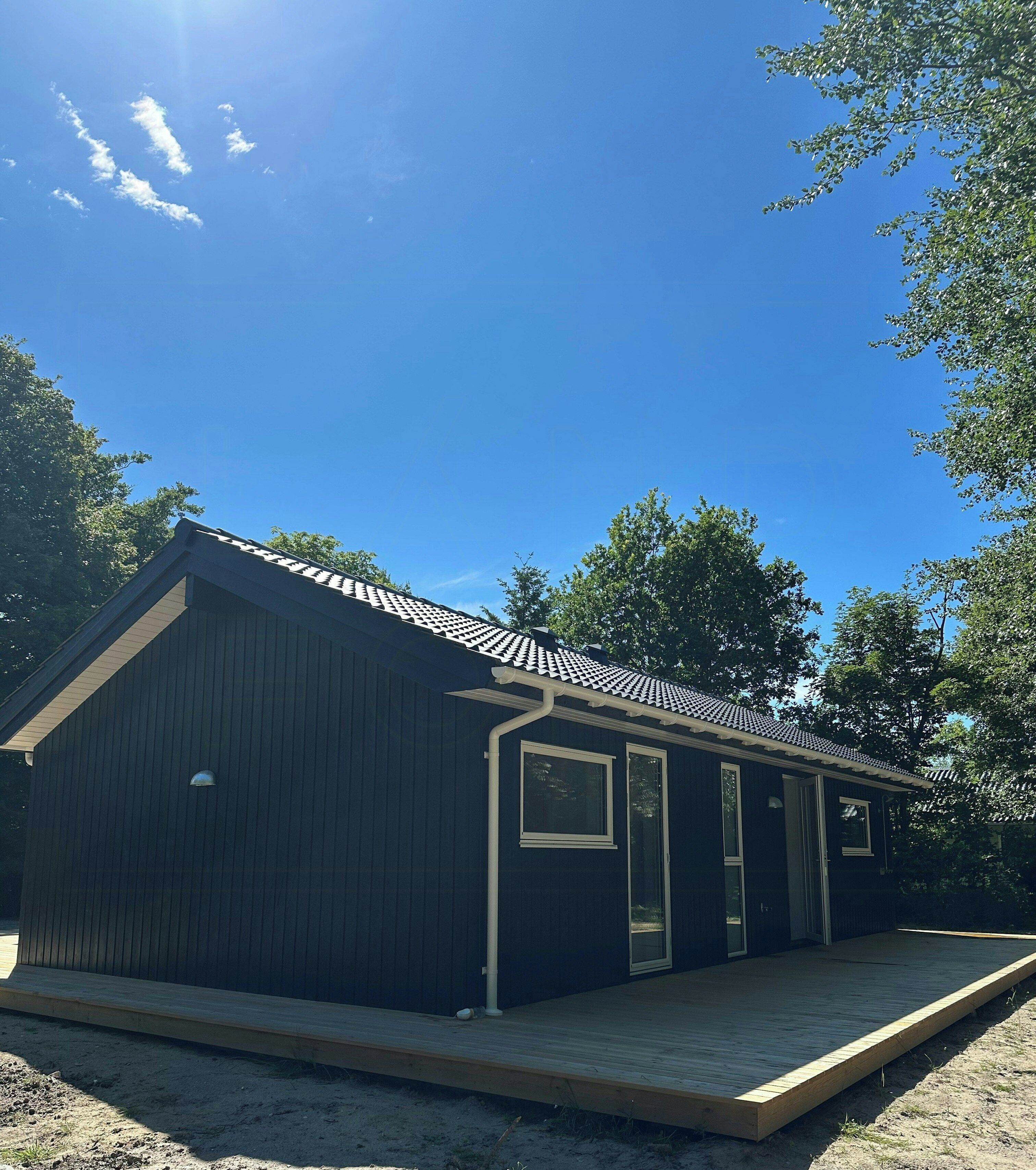 A small, dark-colored building with a sloped roof, a wooden deck, and a clear blue sky.