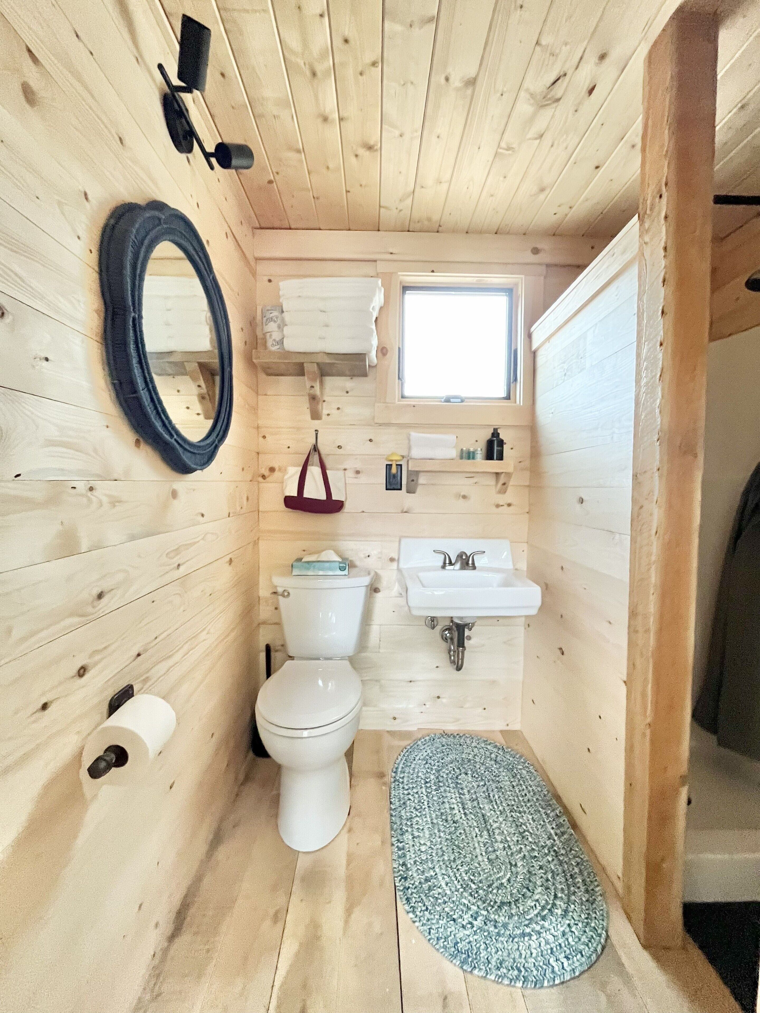 A small, wooden-paneled bathroom with a toilet, sink, mirror, and a window.