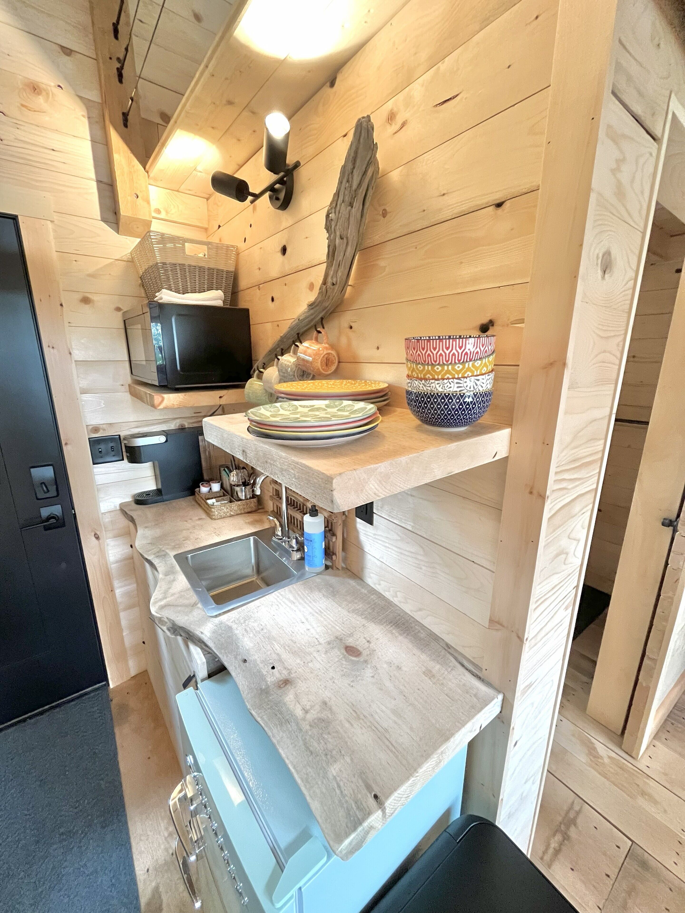 A compact kitchen with wooden walls, a microwave, a sink, and a shelf with bowls.
