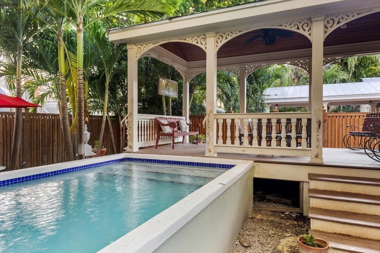 A pool area with a covered seating area, surrounded by a wooden fence and palm trees.