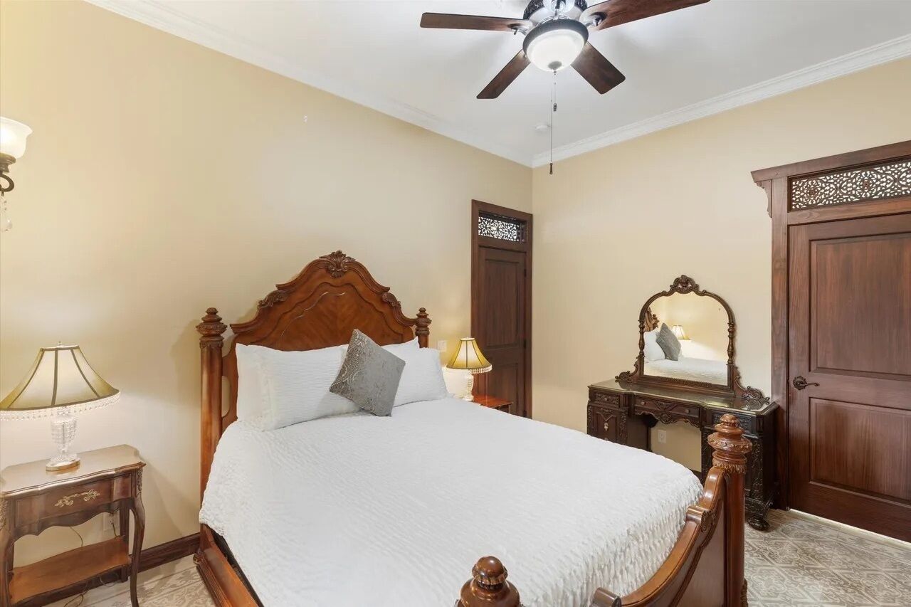 A bedroom with a wooden bed, a ceiling fan, a nightstand with a lamp, and a mirror on a dresser.